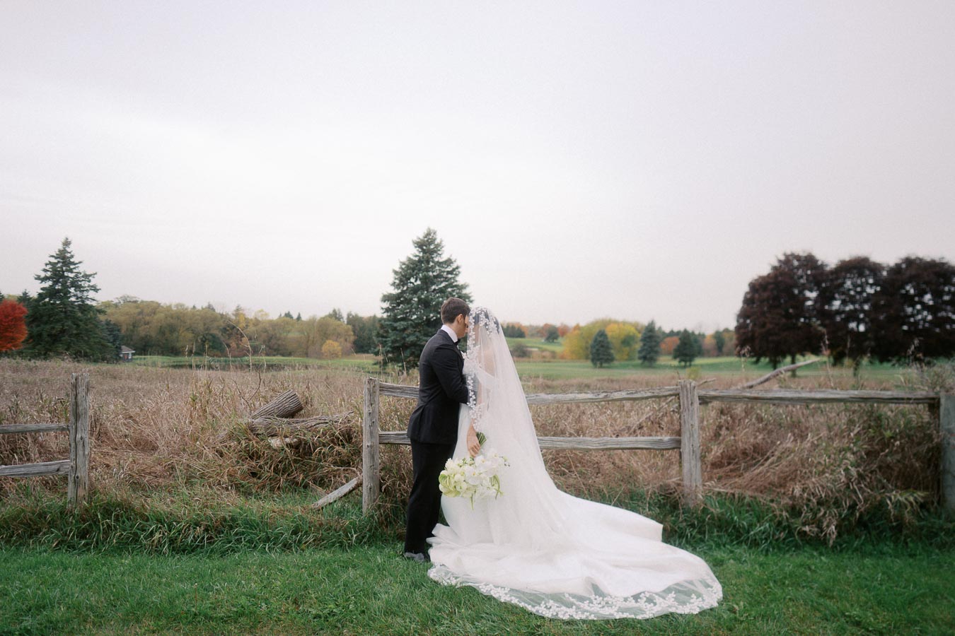 Bride and groom sharing an intimate moment outdoors by a rustic wooden fence, surrounded by lush greenery and autumn trees, bridal gown with a train, wedding bouquet in hand.