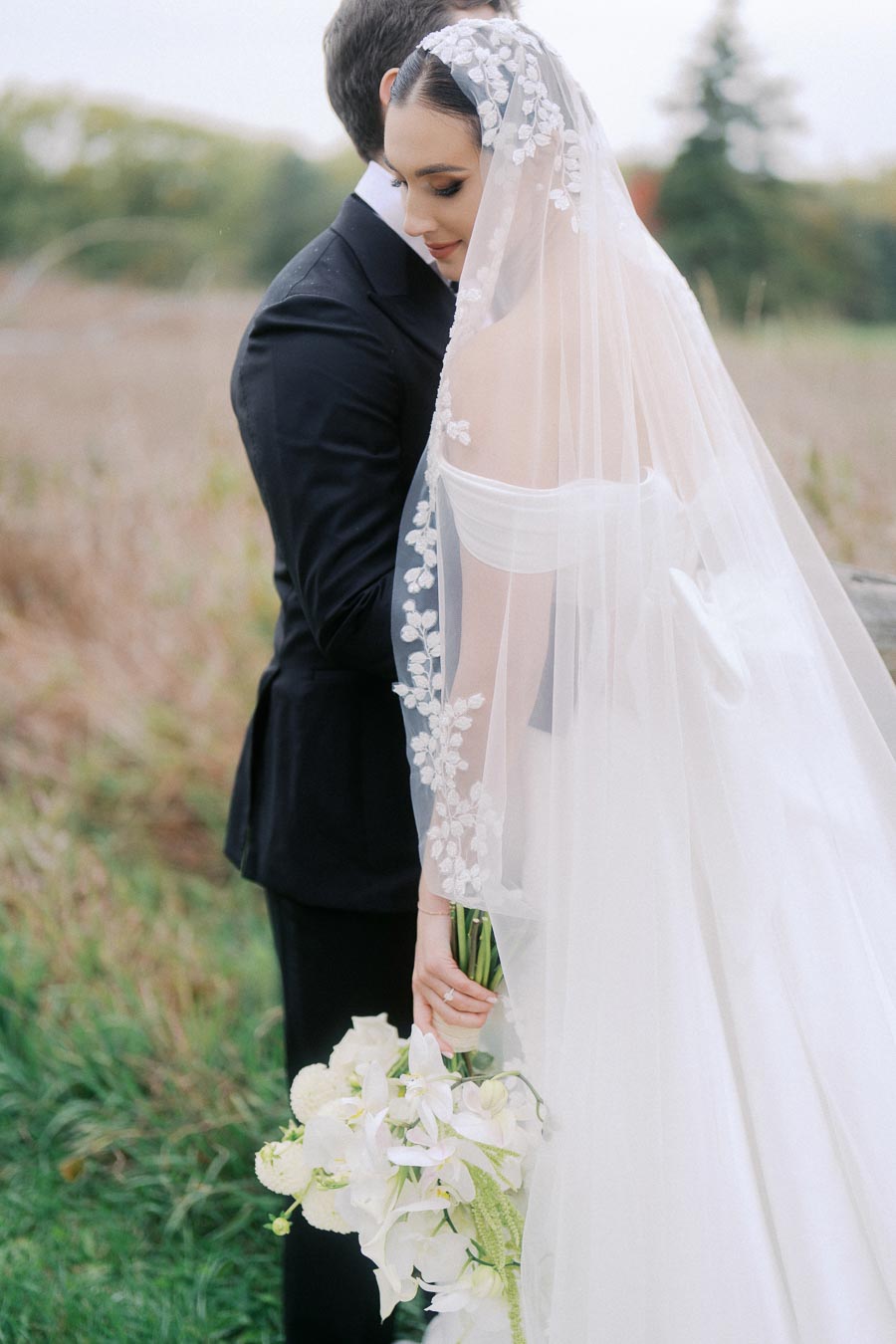 Bride in elegant white wedding gown with floral veil holding bouquet of white flowers, standing in a serene outdoor setting with a groom in a black suit, surrounded by lush greenery.