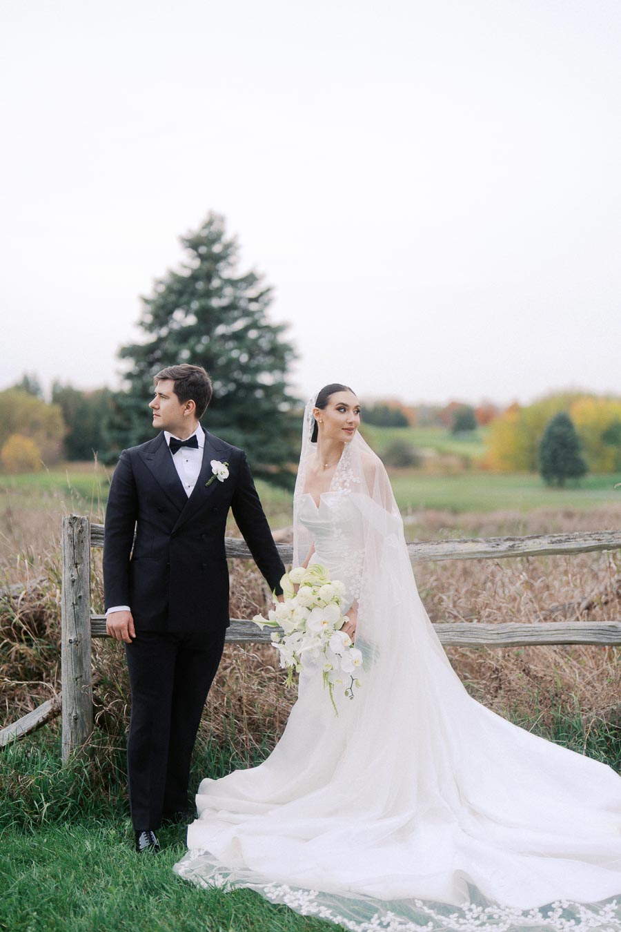 A bride and groom stand together outdoors by a rustic wooden fence, with the bride in a flowing white gown holding a bouquet of white flowers, and the groom in a black tuxedo, set against a backdrop of green fields and trees.