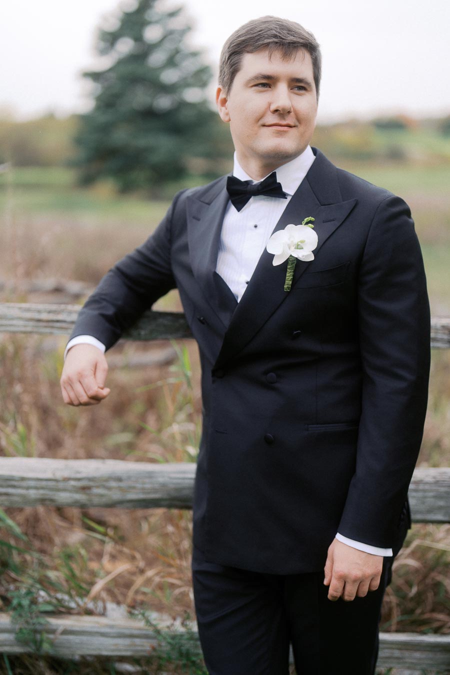 Young man in elegant black tuxedo with white floral boutonniere, standing outdoors near rustic wooden fence, with greenery and blurred background.