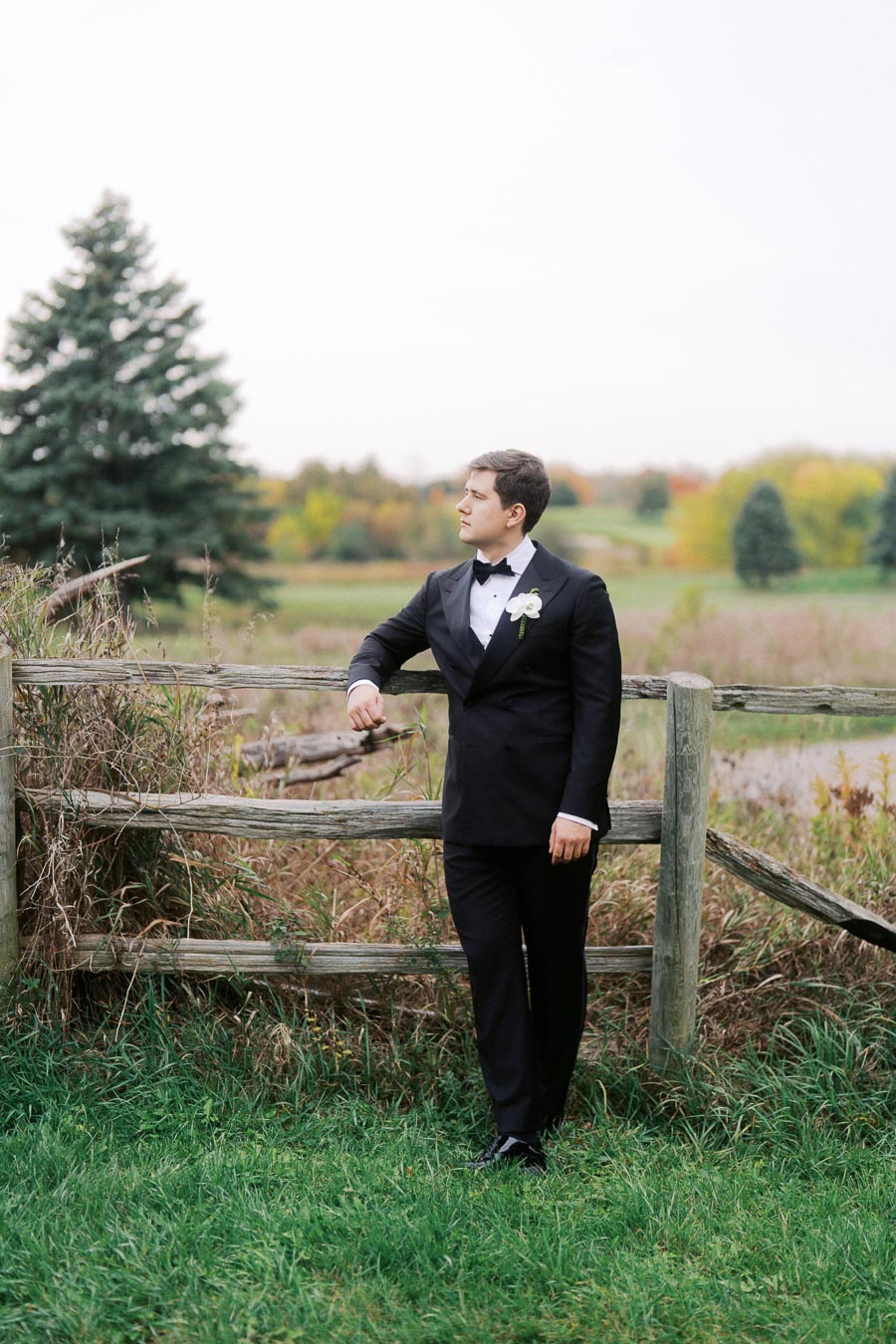 A groom in a black tuxedo looks thoughtfully into the distance while leaning against a rustic wooden fence in a scenic countryside setting with autumn foliage.