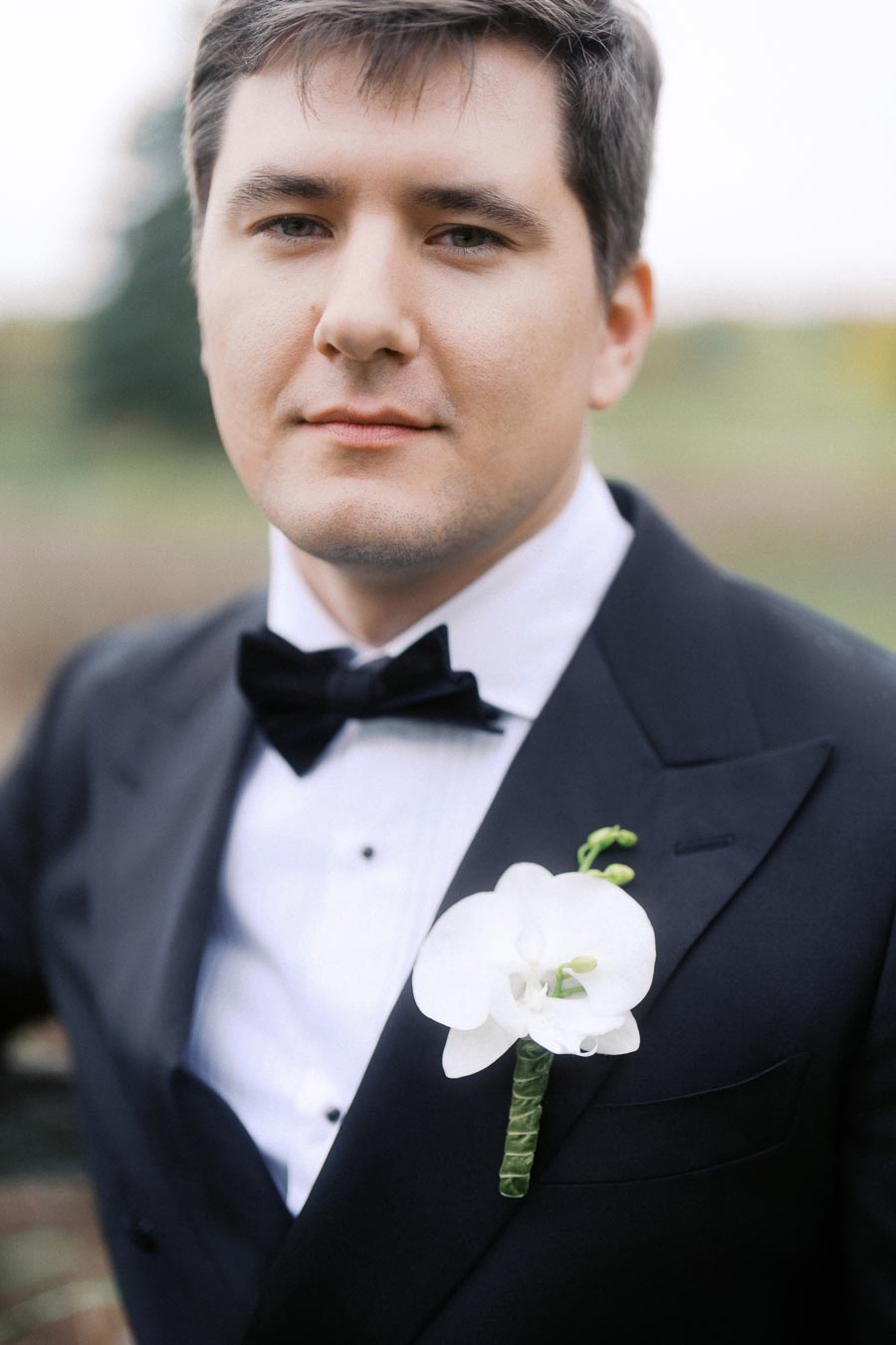 A man dressed in a black tuxedo with a white shirt and black bow tie, wearing a white orchid boutonniere, outdoors.