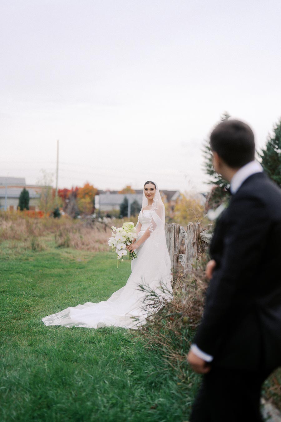 Bride in elegant white wedding dress holding a bouquet, standing near a rustic fence in a scenic outdoor setting, with a groom in the foreground gazing at her.