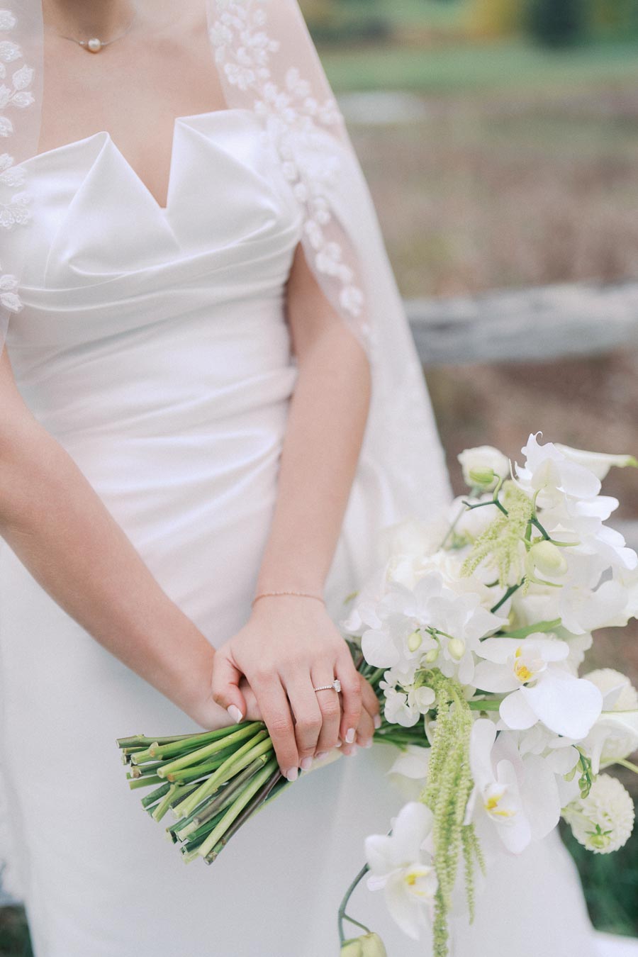 Bride in elegant white gown holding a bouquet of white orchids and greenery, showcasing a delicate wedding ring, ideal for a romantic outdoor wedding setting.