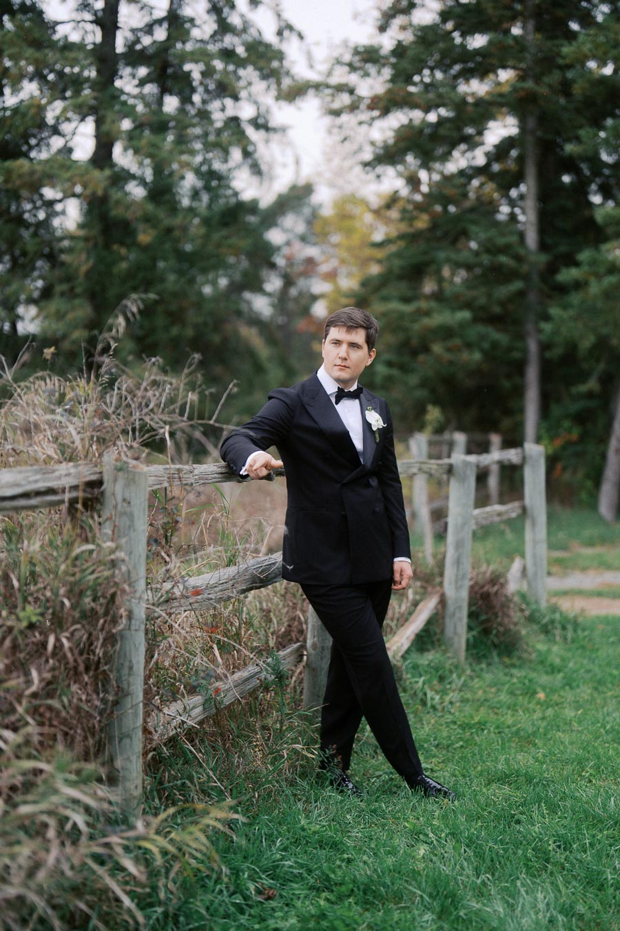 Stylish man in a black tuxedo leaning on a rustic wooden fence in a lush, green outdoor setting.