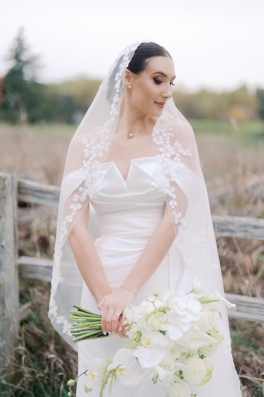 A bride in an elegant white wedding dress and floral veil, holding a bouquet of white flowers, poses outdoors against a rustic wooden fence and natural background.