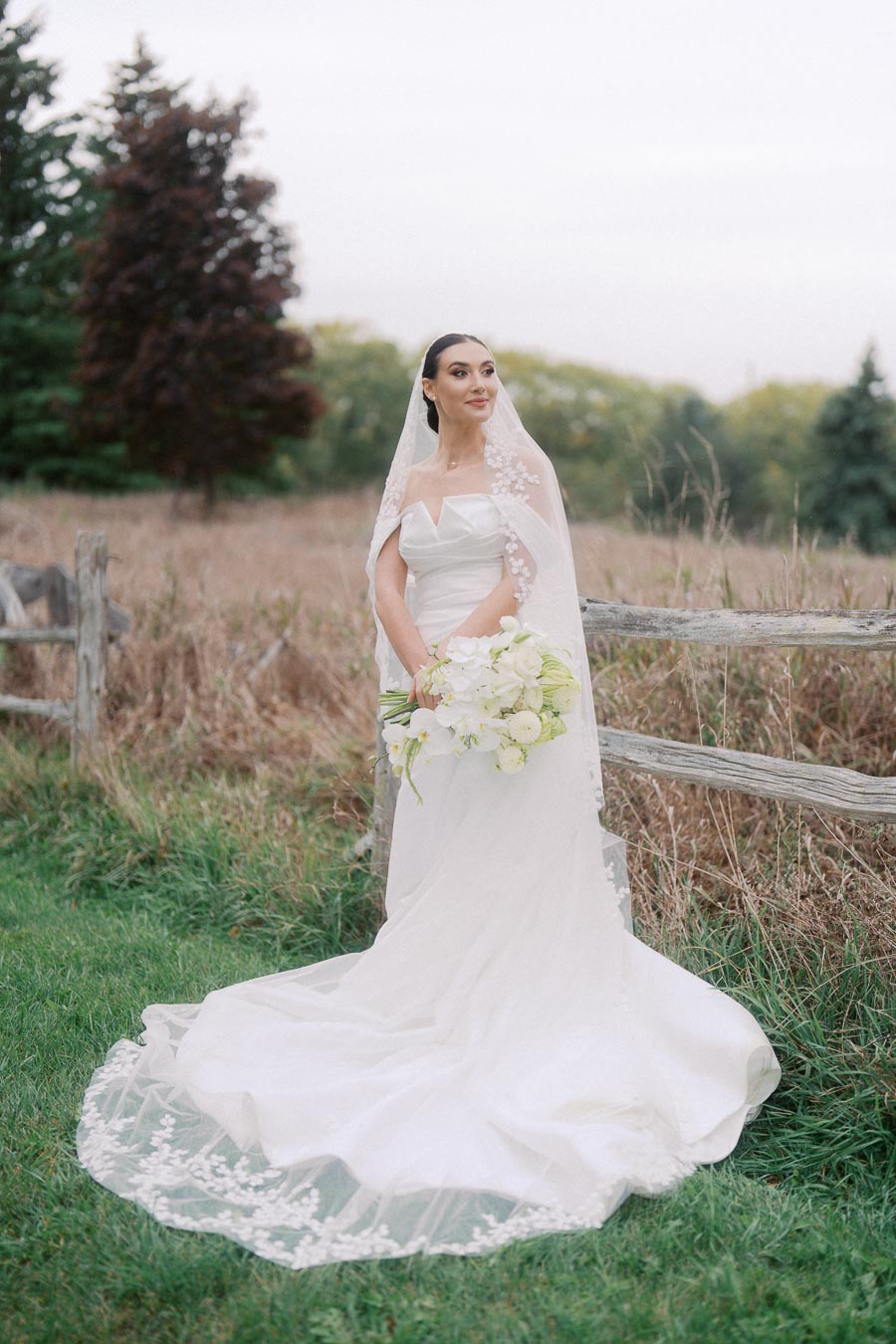 Bride in elegant white gown holding a bouquet of white flowers, standing outdoors near a rustic fence with a natural, forested background.