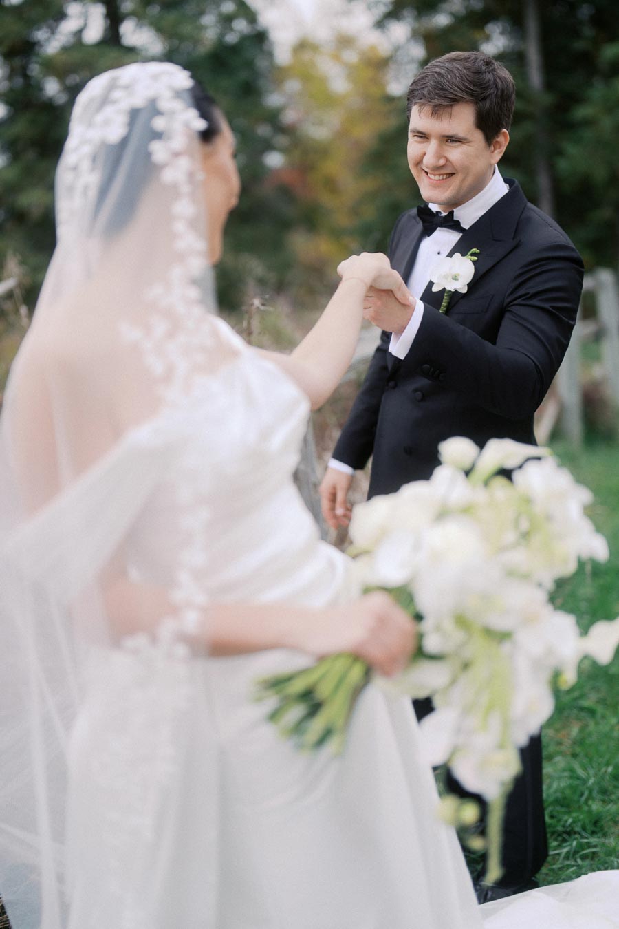 A joyful groom in a black tuxedo holding the hand of a bride in a white wedding gown with floral bouquet, set against a lush outdoor backdrop.