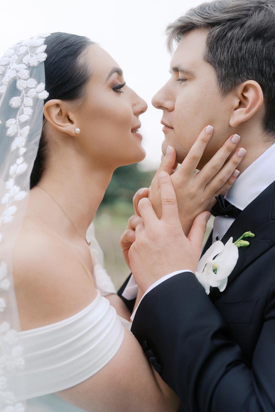 A romantic close-up of a bride and groom gazing into each other's eyes on their wedding day, the bride wearing a delicate lace veil and off-shoulder gown, and the groom in a classic tuxedo with a white floral boutonniere.