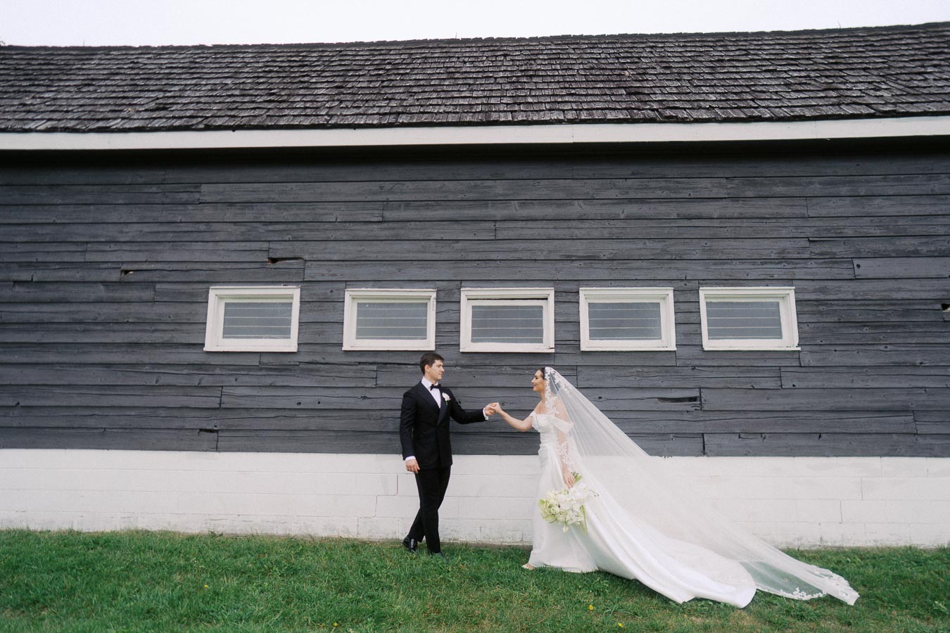 Bride and groom holding hands in front of rustic barn wall, bride wearing elegant white gown with long veil, groom in black tuxedo, outdoor wedding setting.