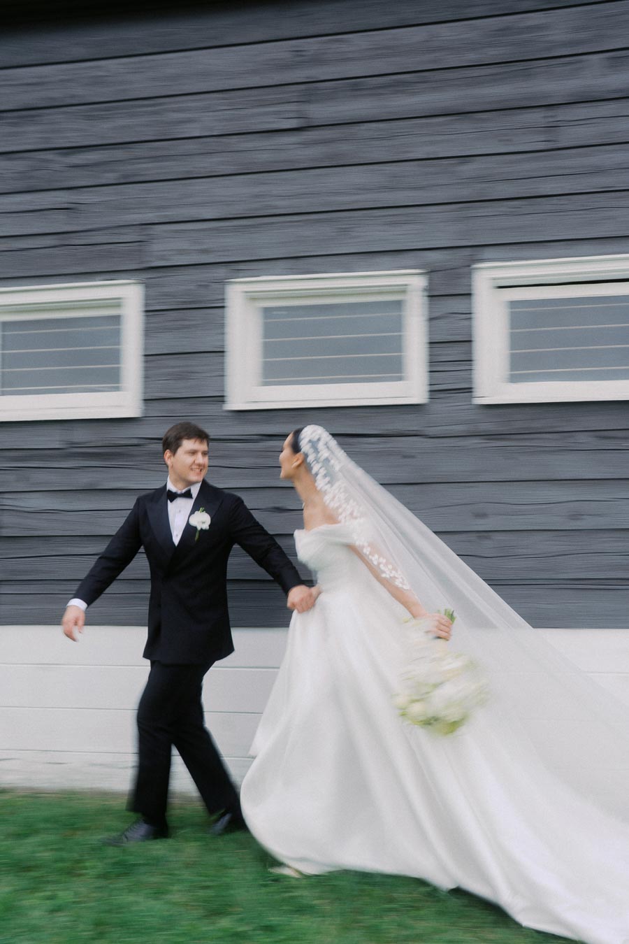 Bride and groom joyfully walking hand in hand, bride wearing a flowing white gown and veil, near a rustic wooden building.