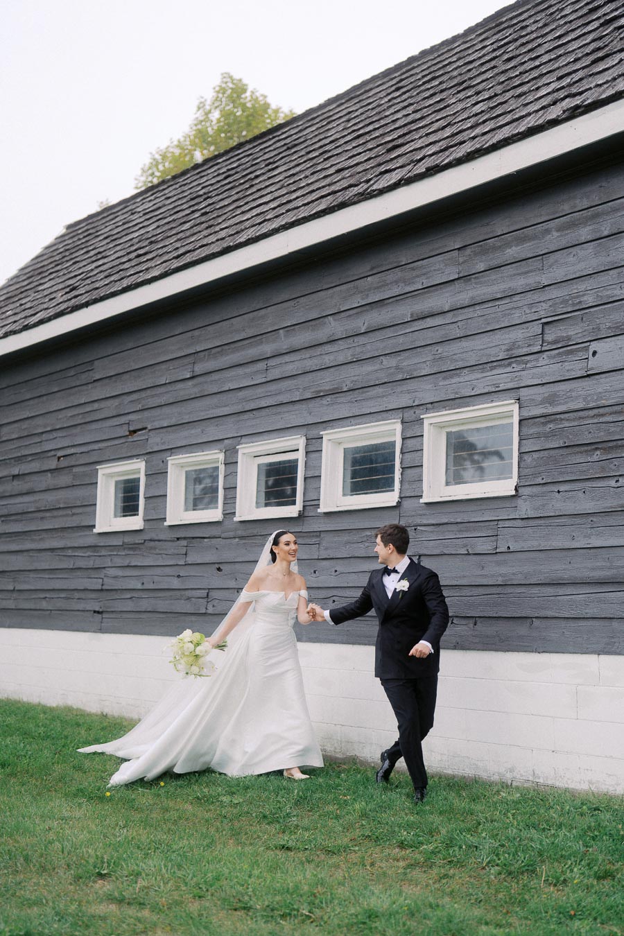 Bride and groom holding hands, walking joyfully outside by a rustic barn. The bride is in a flowing white wedding dress, and the groom is in a black tuxedo, both smiling against a serene backdrop.