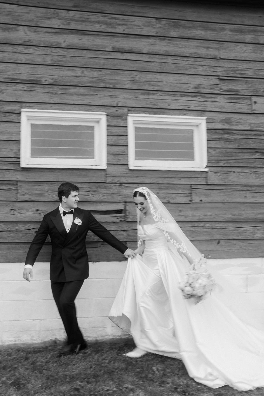Black and white photo of a bride and groom walking hand in hand, with the bride in a classic white wedding dress and veil, and the groom in a tuxedo, set against a rustic wooden barn background.