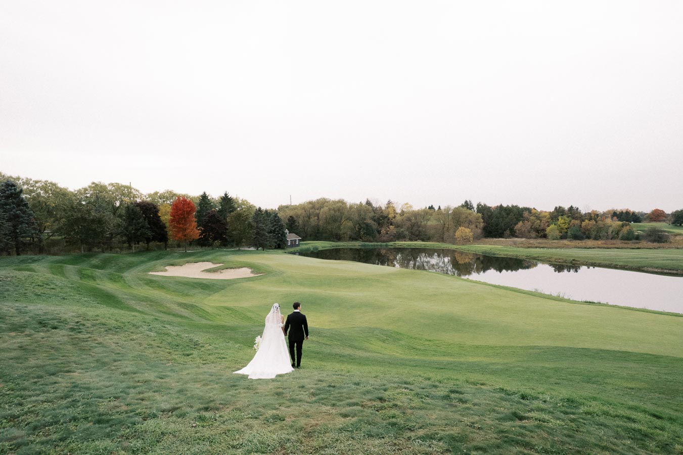 Bride and groom walking on a scenic golf course with a lake and autumn trees in the background, capturing a serene wedding moment.