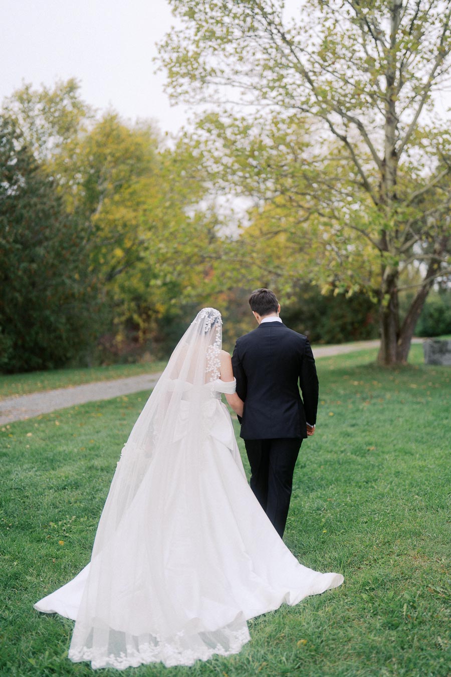 Bride and groom walking away on a lush green lawn against a backdrop of trees, with the bride wearing a white gown and veil, creating a serene and romantic wedding scene.