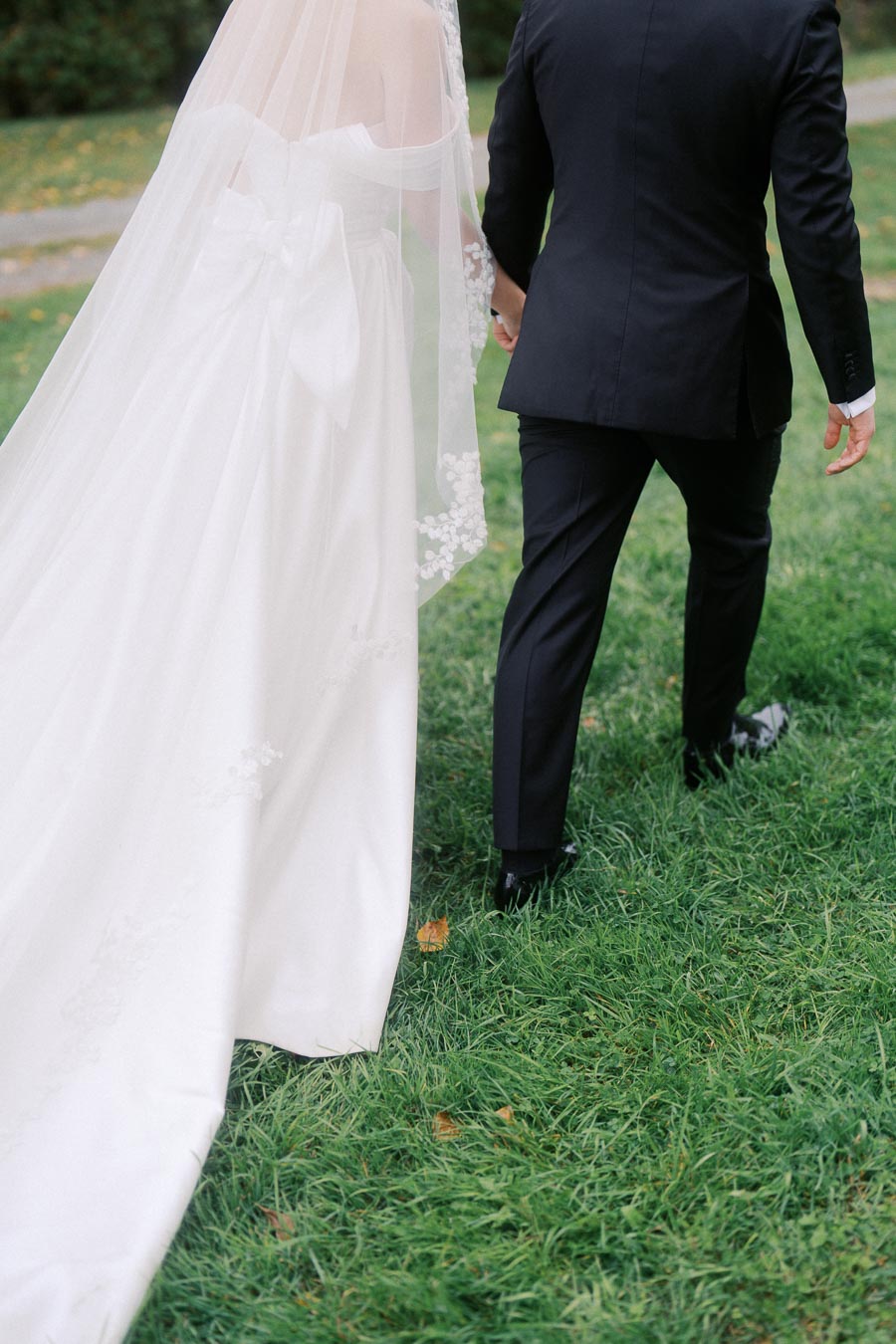Bride and groom walking hand in hand on a lush green lawn, the bride wearing a flowing white wedding dress with a long veil, and the groom in a classic black suit.