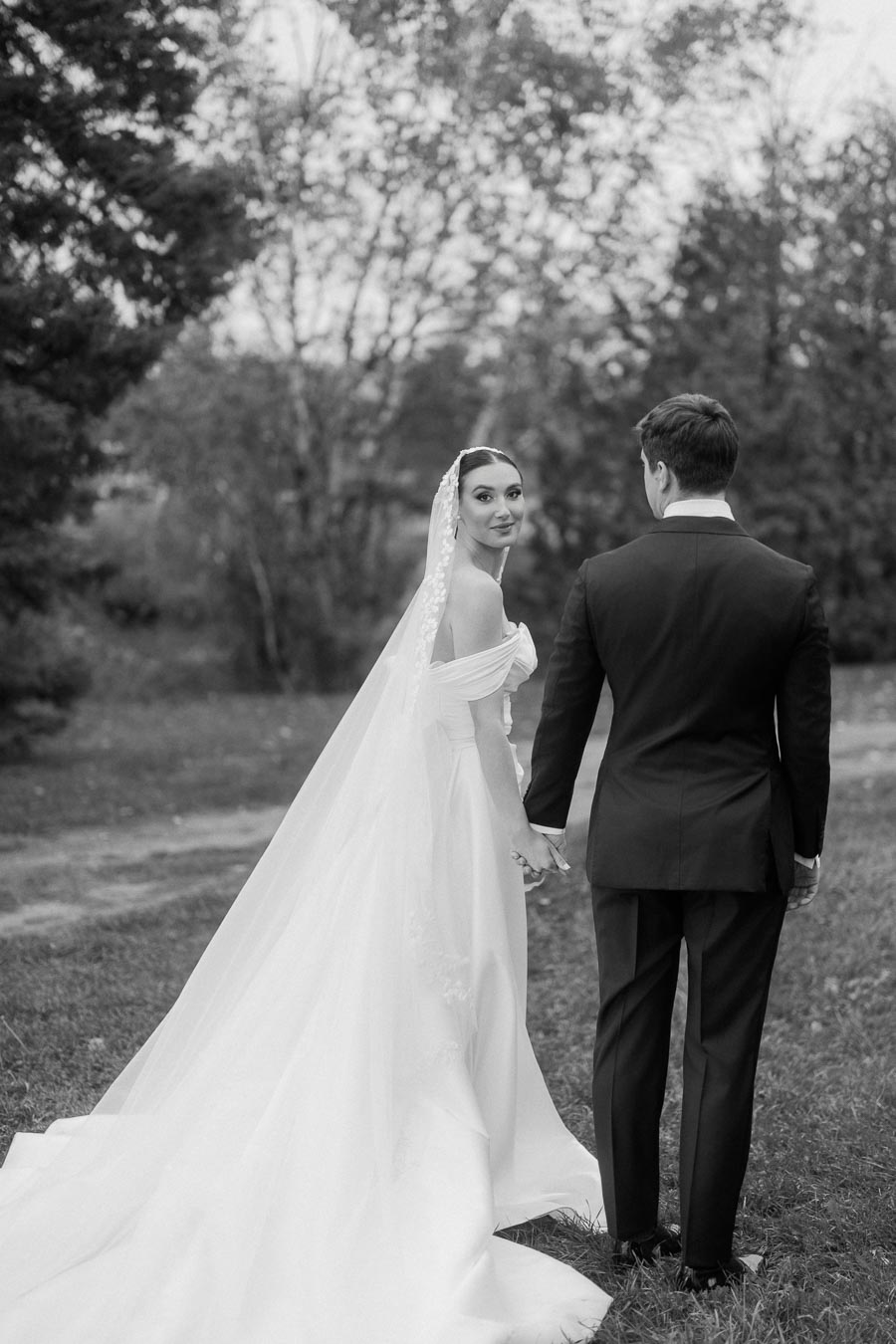 Black and white photo of a bride in an elegant wedding dress with a long train and lace veil, holding hands with her groom in a classic suit, standing outdoors in a scenic, wooded area.