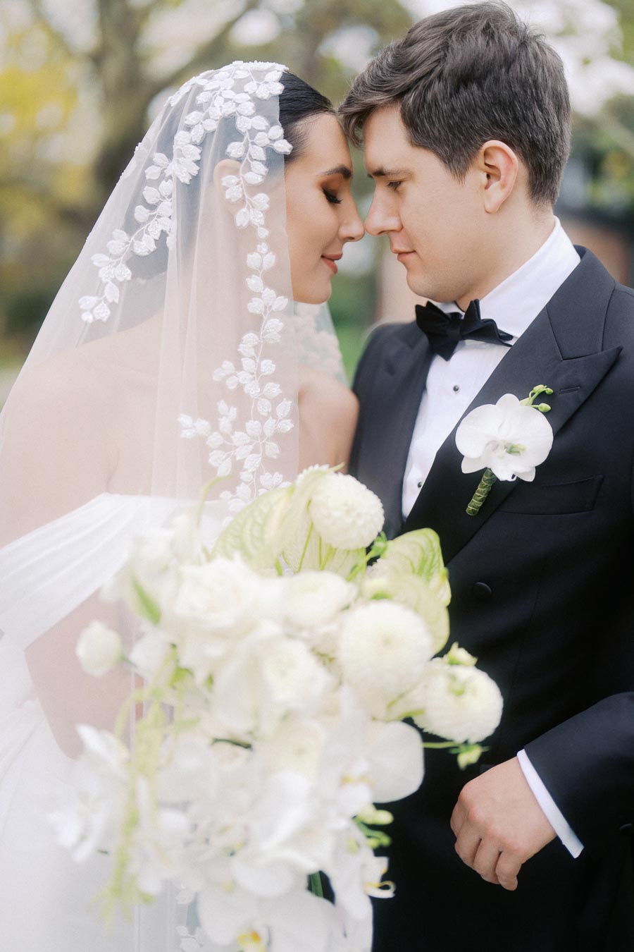 Bride and groom sharing an intimate moment on their wedding day, featuring the bride in an elegant white gown and lace veil, and the groom in a classic tuxedo with a white orchid boutonniere, set against a serene outdoor backdrop.