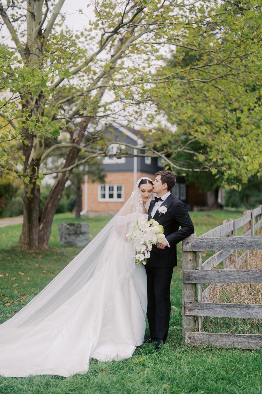 Elegant bride and groom embrace outside by tree and wooden fence, bride in flowing white gown and groom in black tuxedo, holding white and green bouquet near rustic countryside home.
