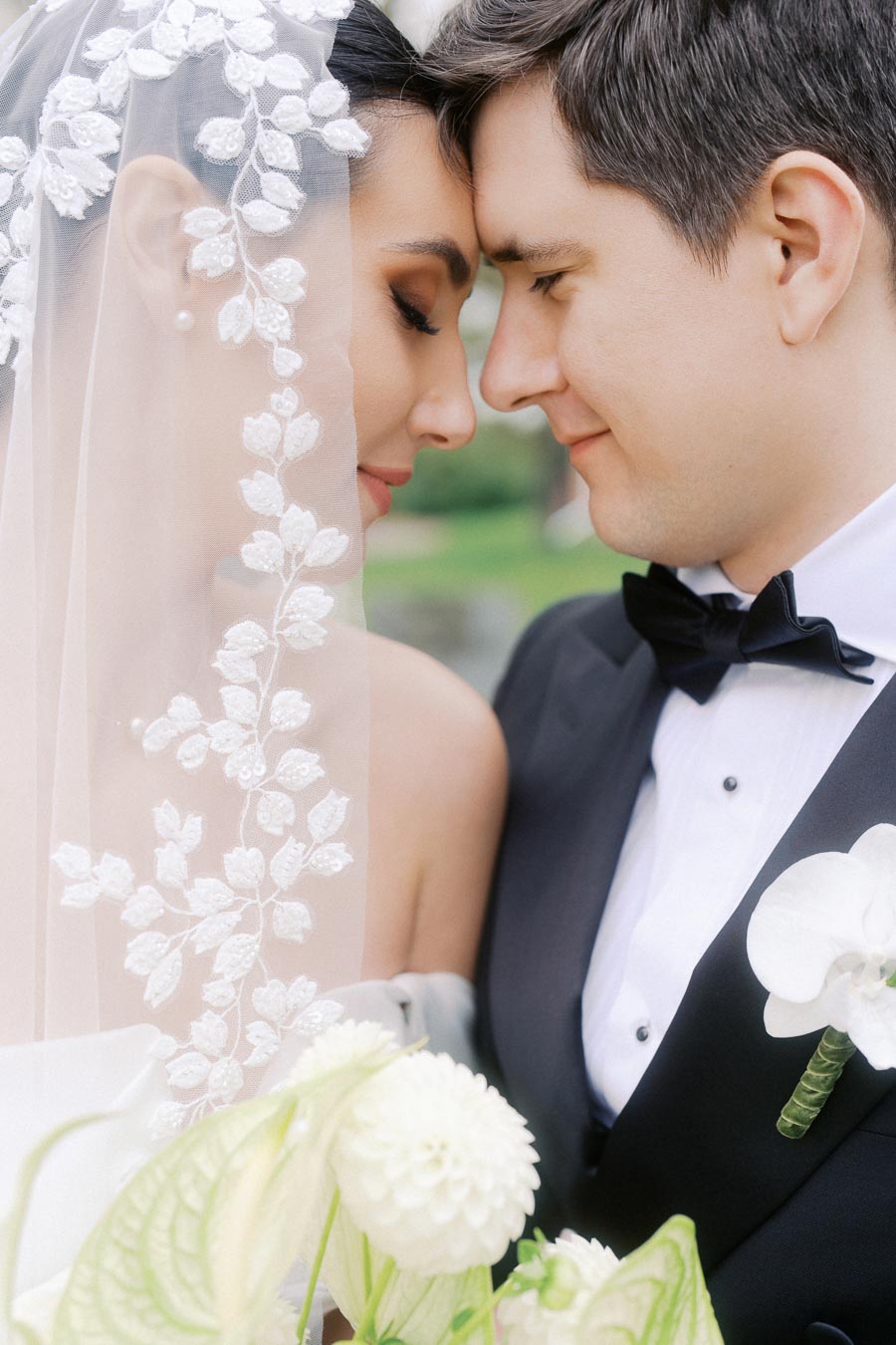 Tender wedding moment with a bride and groom touching foreheads, featuring the bride's elegant lace veil and the groom's classic black tuxedo. Romantic outdoor setting with soft-focus greenery and white flowers in the foreground.