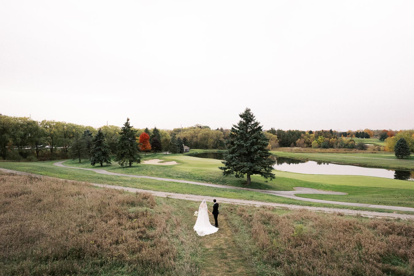 Bride and groom standing in a serene landscape with lush green grass, trees, a pond, and a clear sky, creating a picturesque outdoor wedding setting.
