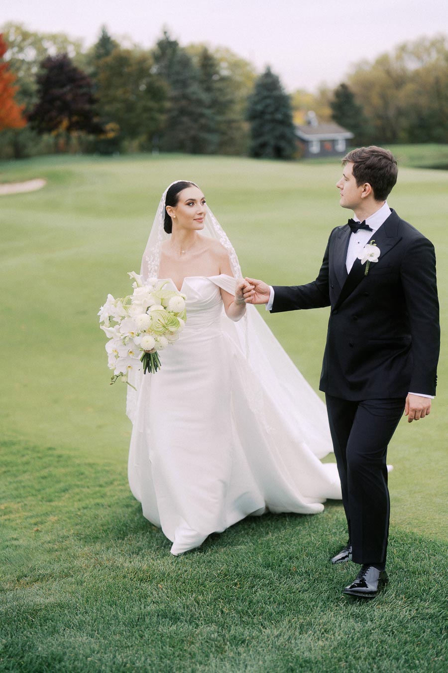 Elegant bride in a white gown holding a bouquet, walking hand in hand with the groom in a black tuxedo on a lush green golf course.