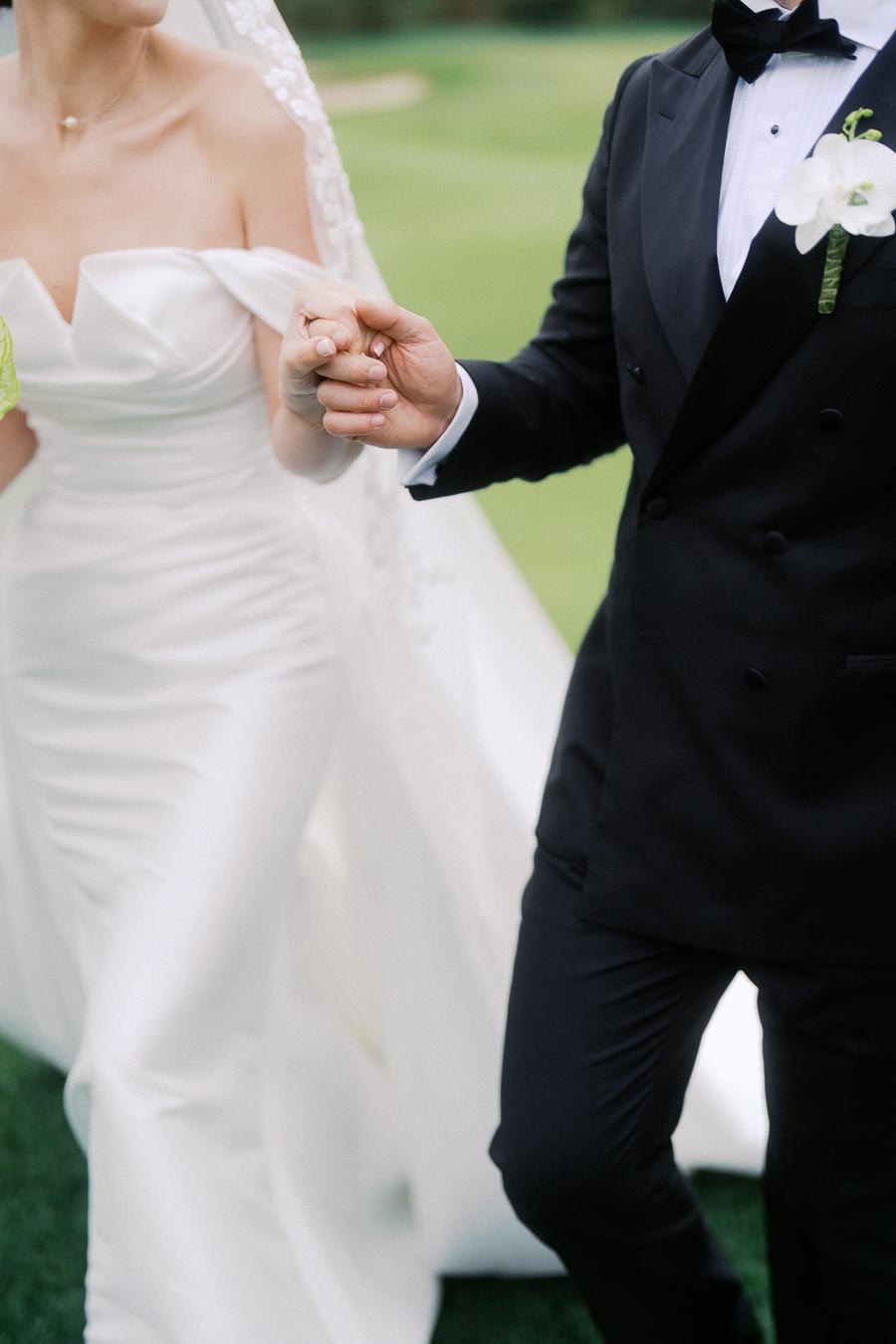 Elegant wedding couple holding hands, with the bride in an off-shoulder white gown and veil, and the groom in a black tuxedo with a boutonniere, standing on lush green grass.