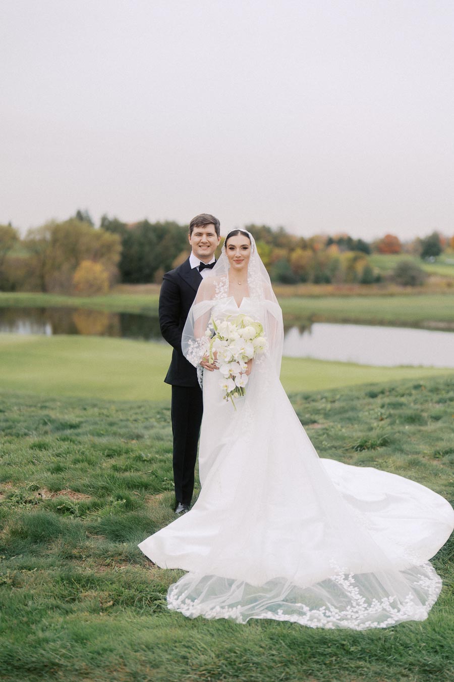 Bride and groom in elegant wedding attire pose outdoors by a scenic pond and lush greenery, showcasing a beautiful wedding scene.
