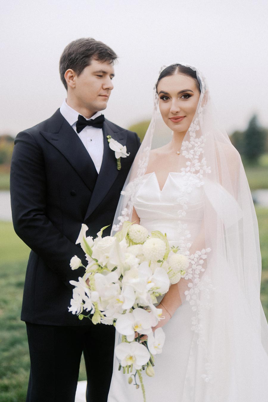 Elegant bride and groom on their wedding day, with the bride in a lace veil holding a bouquet of white flowers, and the groom in a black tuxedo, standing outdoors.