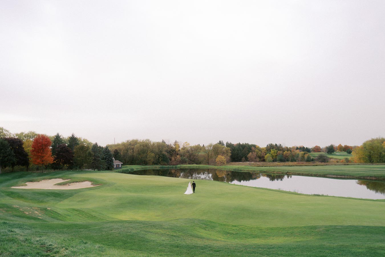 Bride and groom walking hand in hand on a lush green golf course beside a reflective pond, surrounded by autumn trees with vibrant foliage.