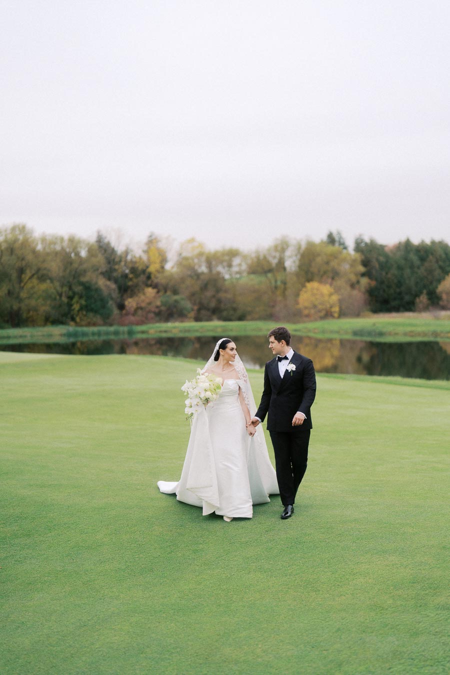 Bride and groom walking hand in hand on a lush green lawn, with a serene backdrop of trees and a small pond, showcasing a beautiful outdoor wedding setting.