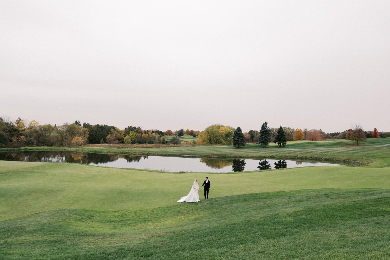 A couple in wedding attire walks hand in hand on a lush green golf course, with a tranquil pond and autumn-colored trees in the background.