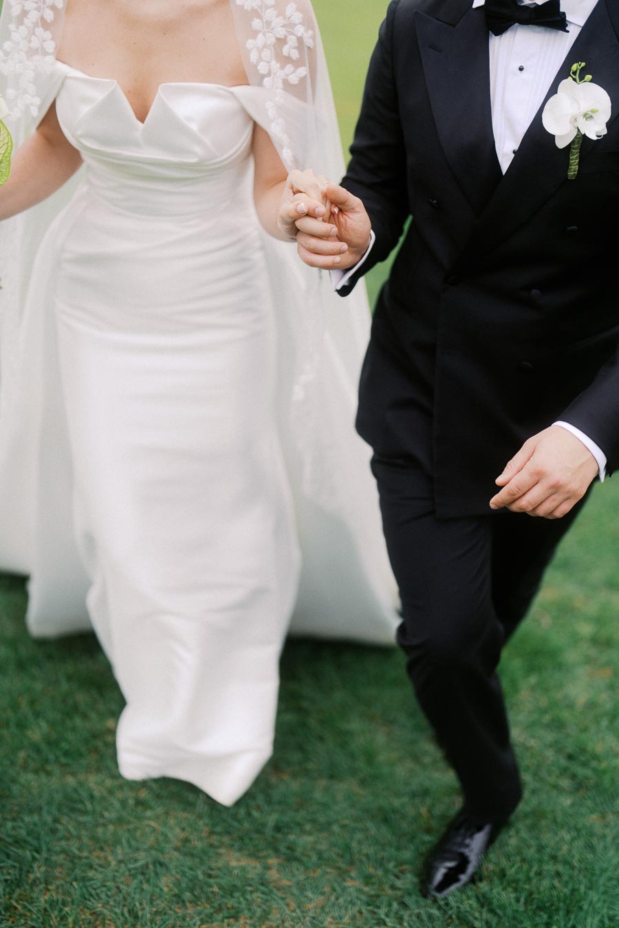 A bride and groom in elegant attire hold hands while walking on a green lawn during a wedding ceremony. The bride wears a white satin gown with a floral lace veil, and the groom is dressed in a classic black tuxedo with an orchid boutonniere.