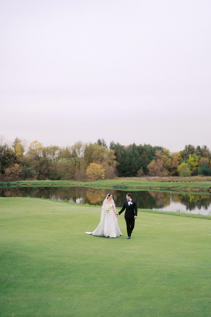 Bride and groom walking hand in hand on a lush green lawn with autumn trees and a serene lake in the background, capturing a romantic outdoor wedding moment.