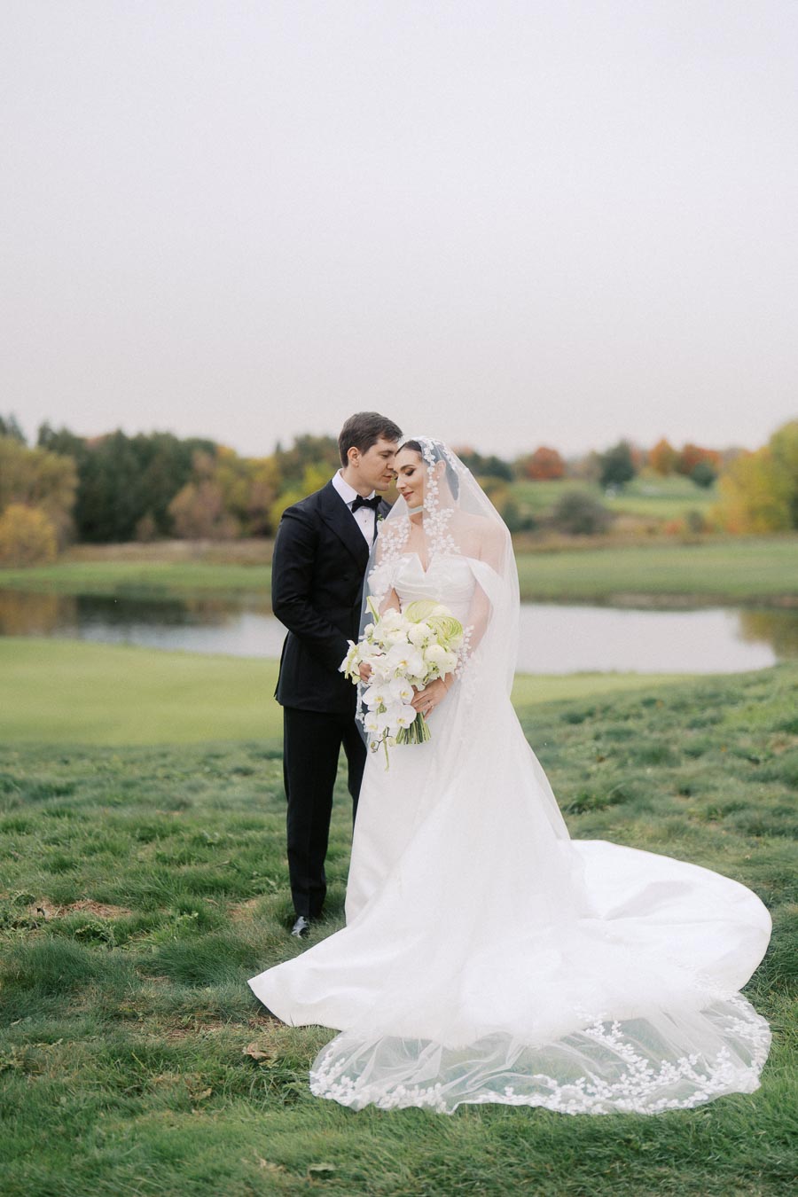 Elegant bride and groom in wedding attire standing on lush green lawn by a scenic pond, with an autumnal backdrop, holding a bouquet of white flowers.