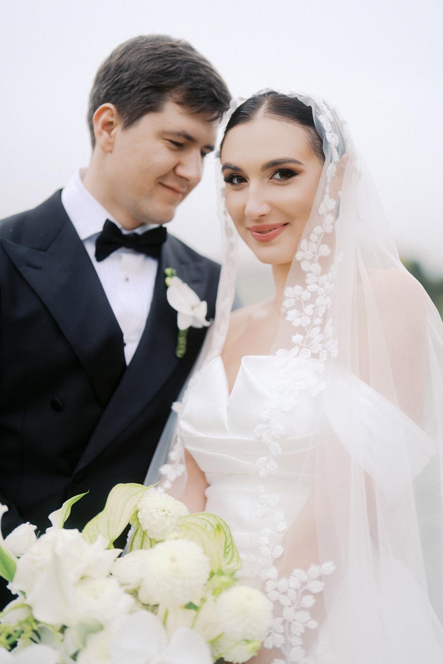 Elegant bride in a floral lace veil and groom in a classic black tuxedo sharing a joyful moment, with the bride holding a bouquet of white flowers.