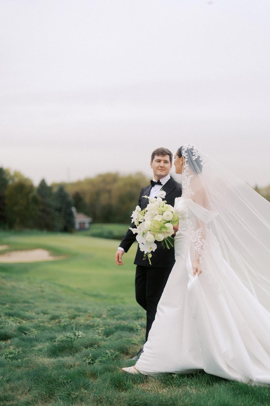Bride and groom walking together outdoors on a lush green lawn, with the bride wearing a white gown and veil, holding a bouquet of white flowers, and the groom in a black tuxedo, capturing a romantic wedding moment.