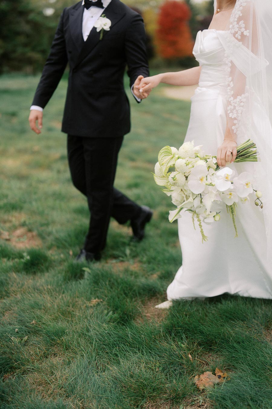 A bride in an elegant white wedding dress holding a bouquet of white flowers, walking hand in hand with a groom in a classic black tuxedo on a grassy lawn.
