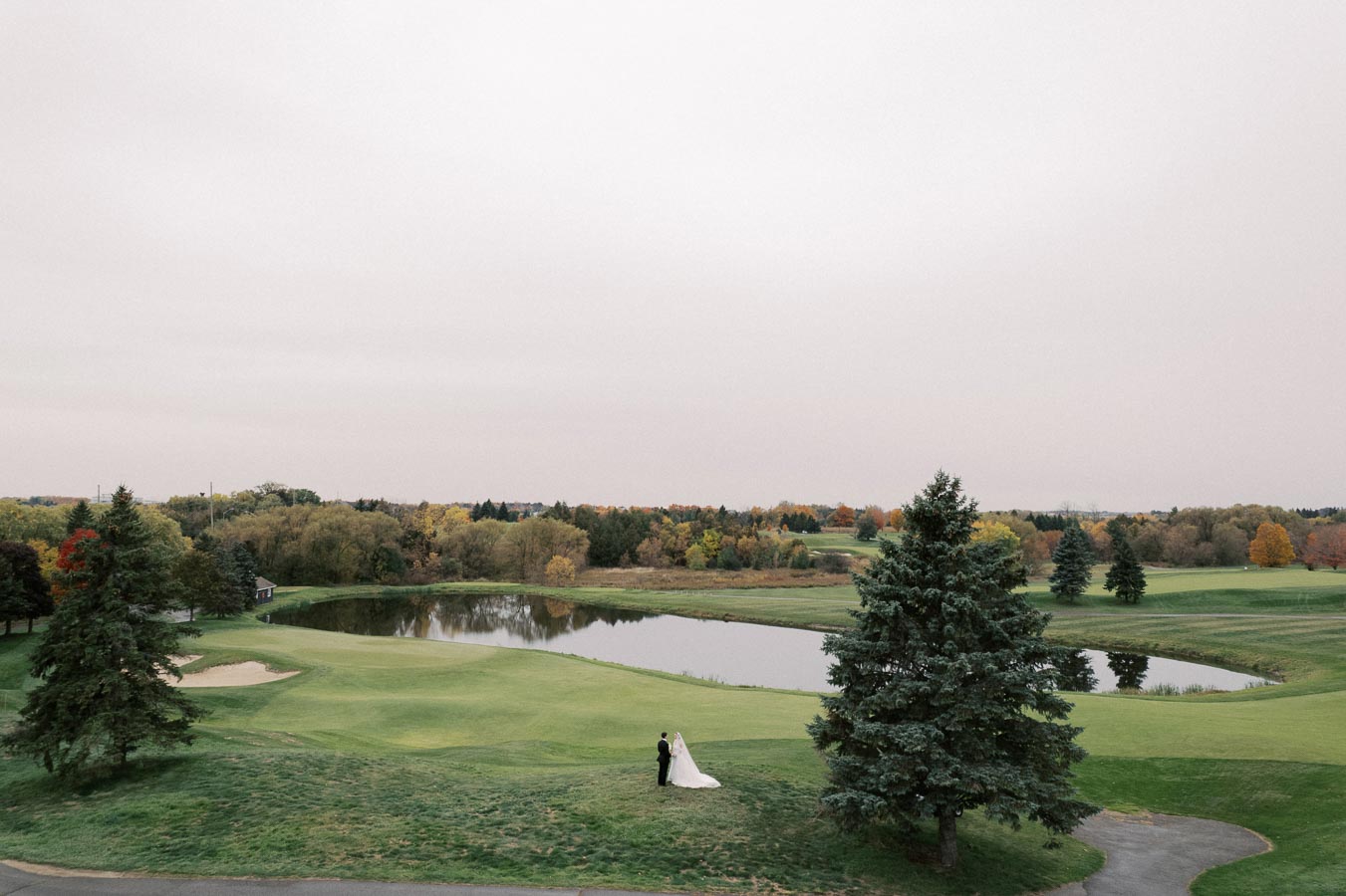 A bride and groom stand on a lush green golf course, surrounded by tall trees with autumn foliage, near a tranquil pond under a cloudy sky.