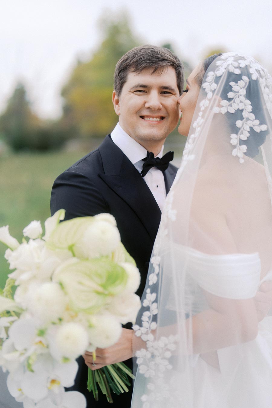 A smiling groom in a black tuxedo holding a bouquet of white flowers, while the bride in a white gown and floral veil kisses his cheek in a lush outdoor setting.