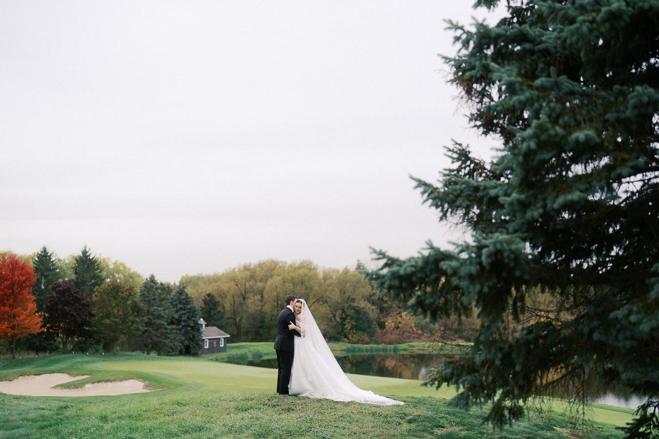 Bride and groom embracing on a scenic golf course, surrounded by lush greenery and autumn foliage, with a serene lake in the background and a large evergreen tree in the foreground.