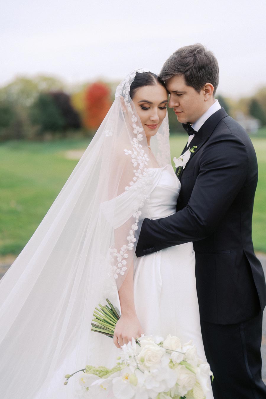 Bride and groom embracing outdoors on their wedding day, with the bride wearing a white dress and veil adorned with floral patterns, holding a bouquet of white flowers, against a backdrop of lush greenery and autumn colors.