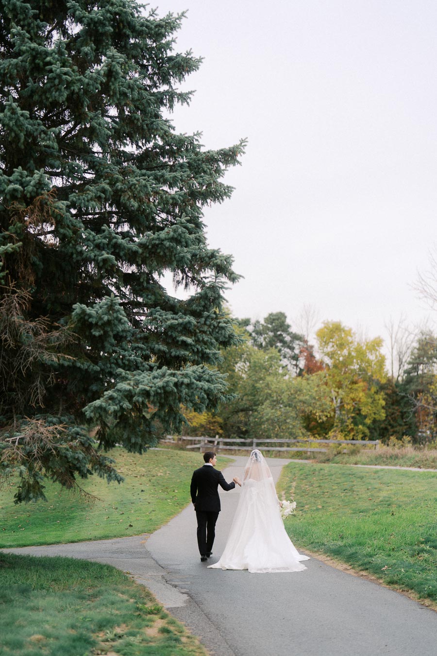 Bride and groom walking along a scenic path surrounded by lush greenery and trees, with the bride wearing a flowing white wedding gown and veil, showcasing a romantic outdoor wedding setting.