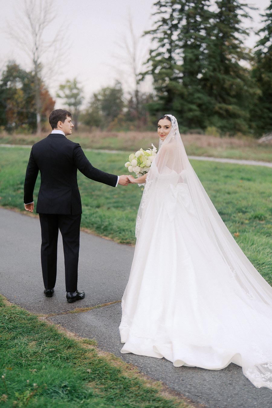 Bride in elegant white wedding gown with long veil holds bouquet of white flowers while looking back, standing on a path in a scenic outdoor setting with groom in black suit; captures romantic wedding moment.