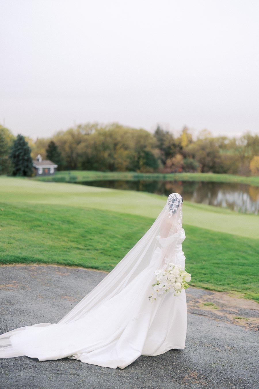 Bride in elegant white wedding gown and lace veil holding a bouquet of white flowers, standing on a golf course near a serene pond with lush greenery in the background.