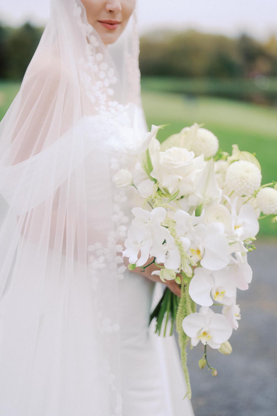 Bride in an elegant white dress holding a bouquet of white flowers and orchids, partially veiled, standing on a picturesque outdoor setting.
