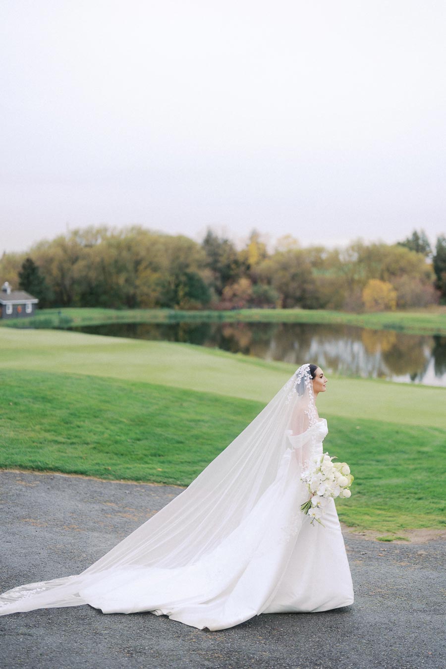 Bride in white wedding gown with long veil walking by a serene lake and lush greenery in a park setting.