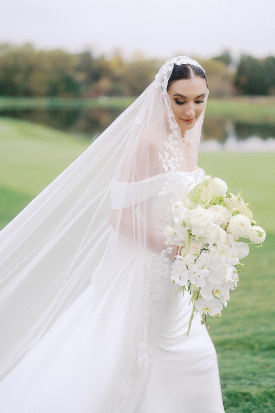 Bride in elegant white wedding gown with lace veil and floral bouquet, standing outdoors on green lawn.