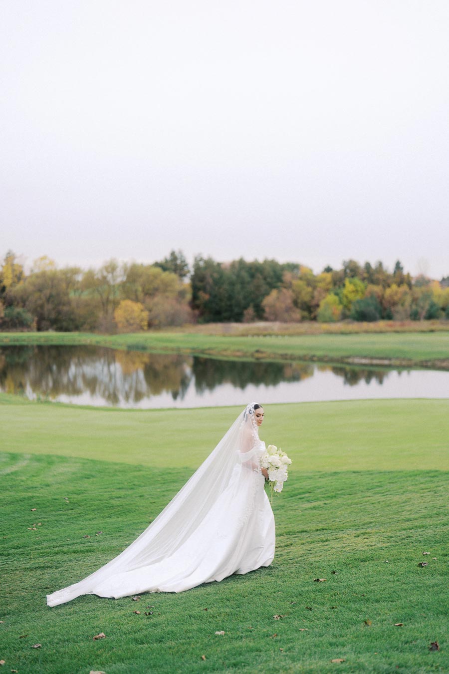 Bride in elegant white wedding dress with long veil holding bouquet, standing on lush green lawn by a serene lake with autumn trees in the background.