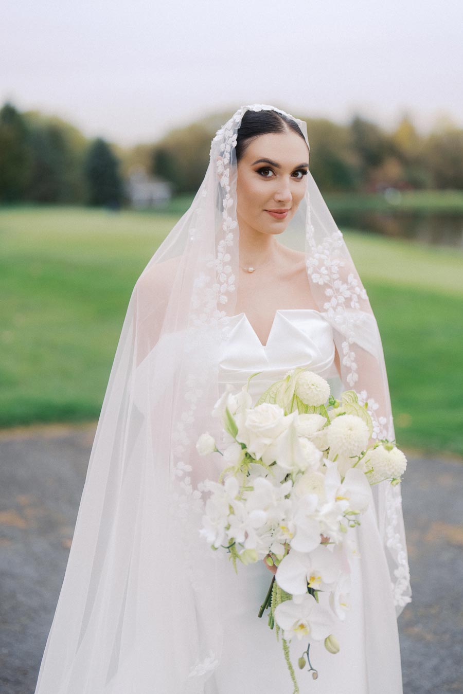 Elegant bride in a white dress with a floral embroidered veil, holding a lush bouquet of white flowers, standing outdoors on a scenic lawn.